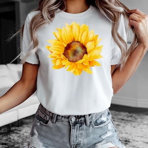 May include: White t-shirt featuring a large, vibrant watercolor sunflower. The sunflower has bright yellow petals and a brown center. The shirt is short-sleeved and appears to be made of a soft material. The model is wearing denim shorts.