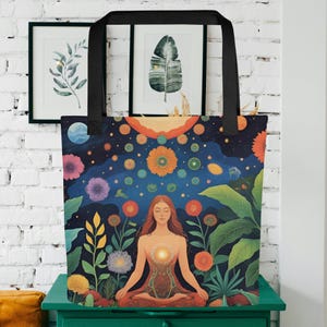 May include: A tote bag featuring a vibrant illustration of a person meditating amidst a celestial and floral landscape. The design includes a night sky with stars, flowers, and a sun, with a person in a meditative pose. The bag has black handles.