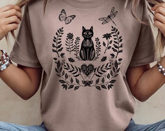 May include: A tan t-shirt featuring a black cat design surrounded by floral and butterfly illustrations. The cat has intricate patterns, and the design includes a heart-shaped element. The shirt is a casual wear item.
