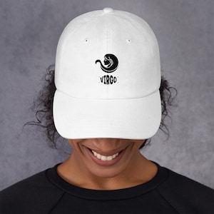 May include: A white baseball cap featuring a black Virgo zodiac symbol and text. The cap has a curved brim and a classic six-panel design. The Virgo symbol is a stylized woman's head with flowing hair.