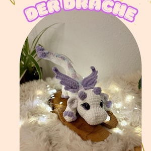 May include: A handmade white and lavender crochet dragon with purple wings and horns. The dragon is displayed on a wooden board atop a fluffy white surface. Text in German reads "LIORA DER DRACHE".
