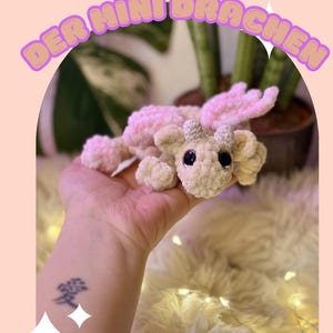 Puede incluir: Un dragón de ganchillo hecho a mano en crema y rosa, con ojos negros grandes y pequeños cuernos. El dragón está sobre una superficie suave de color crema. La imagen incluye el texto "ELMI DER MINI DRACHEN".