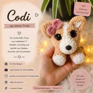Könnte beinhalten: Ein gehäkeltes Corgi-Stofftier aus beigem und weißem Garn mit einer rosa Schleife auf dem Kopf und großen dunklen Augen. Das Bild enthält Text, darunter den Titel Codi der kleine Corgi, und weist auf eine PDF-Anleitung zum Herunterladen hin.