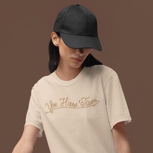 May include: A person wearing a black baseball cap and a cream-colored t-shirt. The t-shirt features the text "Yee Haw'Tism" in a cursive, gold-colored font. The background is a solid brown.