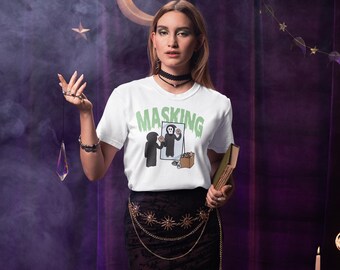 Può includere: Una maglietta bianca con la scritta "MASKING" in lettere verdi. La maglietta presenta un'immagine a cartoni animati di una figura che si guarda allo specchio. La persona indossa un choker nero e una cintura a catena dorata. Lo sfondo è una tenda viola con stelle e una luna crescente.