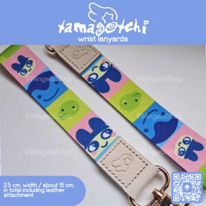 May include: Two wrist lanyards with colorful cartoon character patterns. The lanyards are 2.5 cm wide and about 15 cm long, including the leather attachment. The text "tamagotchi wrist lanyards" is at the top.