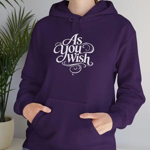 May include: A deep purple hoodie with a front pocket and a drawstring hood. The hoodie features the white text "As You Wish" in an elegant, cursive font. The sleeves are long, and the person is wearing blue jeans.