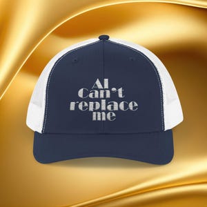 May include: A navy blue trucker hat with a white mesh back. The hat features the embroidered text "AI Can't replace me" in a silver color. The hat has a curved brim and a snapback closure.