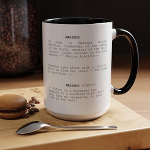 May include: White ceramic mug with a black interior and handle. The mug features text from the movie "Gladiator" about Maximus. A spoon and a macaron are on a wooden surface next to the mug.