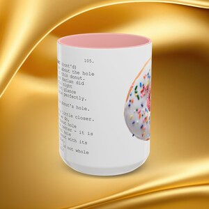 K&ouml;nnte beinhalten: Wei&szlig;e Keramiktasse mit rosa Innenraum, mit einem Teilbild eines Donuts mit wei&szlig;em Zuckerguss und bunten Streuseln. Auf der Seite der Tasse ist Text aufgedruckt. Die Tasse steht vor einem goldenen Hintergrund.