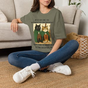 May include: An olive green short-sleeve t-shirt featuring a graphic of two cats wearing green cloaks and vests. The text reads: WE'VE HAD ONE, YES, WHAT ABOUT SECOND BREAKFAST? and I DON'T THINK HE KNOWS ABOUT SECOND BREAKFAST, PIP.