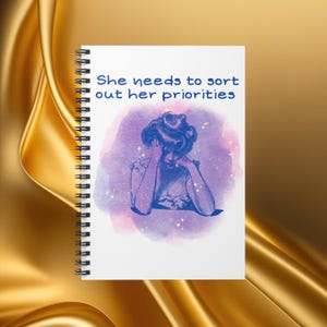May include: A spiral-bound notebook with the text "She needs to sort out her priorities." The cover features a blue illustration of a woman with her head in her hands, set against a purple and pink watercolor background.