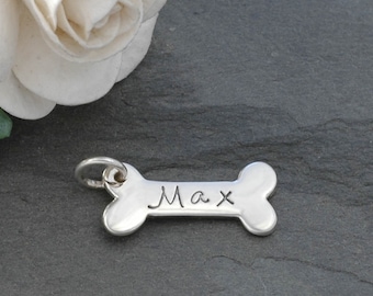 May include: Silver bone-shaped charm with the name Max engraved on it. The charm is sitting on a gray surface with a white rose in the background.