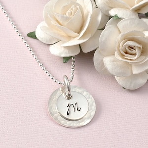 May include: A silver necklace with a hammered silver disc pendant featuring the letter 'm' in cursive script.