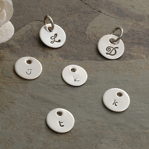 May include: Six silver charms with different letters engraved on them. The letters are J, K, L, D, K, and T.