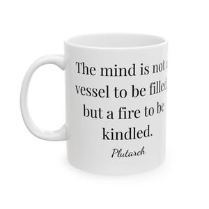 May include: White ceramic mug with a curved handle. The mug features the quote "The mind is not a vessel to be filled but a fire to be kindled" attributed to Plutarch in black text.