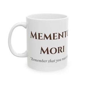 May include: White ceramic mug with the words "Memento Mori" in brown lettering. Below the words is the phrase "Remember that you must die." The mug has a curved handle and a smooth, glossy finish.