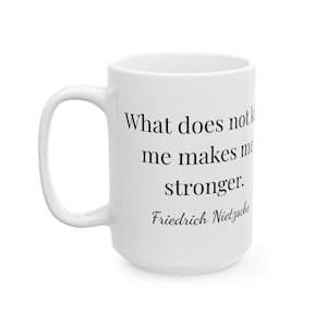 Puede incluir: Taza de cerámica blanca con asa curva. La taza presenta la cita "Lo que no me mata me hace más fuerte" atribuida a Friedrich Nietzsche en texto negro.