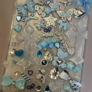 Könnte beinhalten: Eine transparente Tablet-Hülle, verziert mit einer Vielzahl dekorativer Verzierungen. Das Design umfasst hellblaue Schleifen, Schmetterlinge, Herzen, Sterne und andere glitzernde Charms. Ein Hundert-Dollar-Schein ist ebenfalls sichtbar.