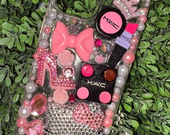 May include: Clear phone case decorated with pink and silver rhinestones, pearls, and charms. Features a pink bow, high heel, crown, and a Hello Kitty design. Includes MAC makeup compact, lipstick, and blush.