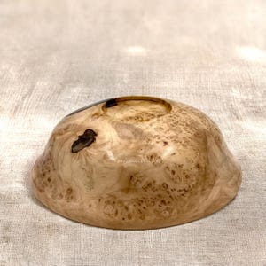 Puede incluir: Un cuenco de madera poco profundo y redondeado con una superficie pulida de color tostado claro, que presenta intrincados patrones de vetas en espiral y nudos naturales de color marr&oacute;n oscuro. Tiene un acabado liso y brillante.