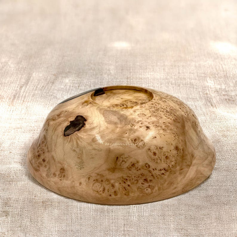 Puede incluir: Un cuenco de madera poco profundo y redondeado con una superficie pulida de color tostado claro, que presenta intrincados patrones de vetas en espiral y nudos naturales de color marr&oacute;n oscuro. Tiene un acabado liso y brillante.