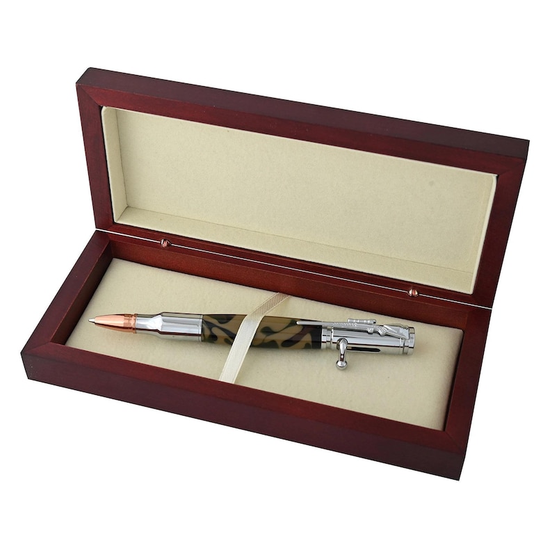 May include: A pen designed to resemble a rifle bullet, presented in a dark wood box with a cream-colored interior. The pen features a copper-colored tip, a camouflage-patterned barrel, and silver-colored metal accents. A cream ribbon secures the pen.