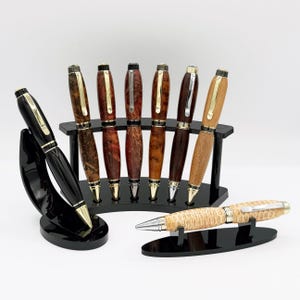 May include: A selection of handcrafted pens with diverse wood grains and finishes, presented on black stands. The pens have gold and silver details, displaying a spectrum of colours from dark brown to light tan. One black pen is also shown.