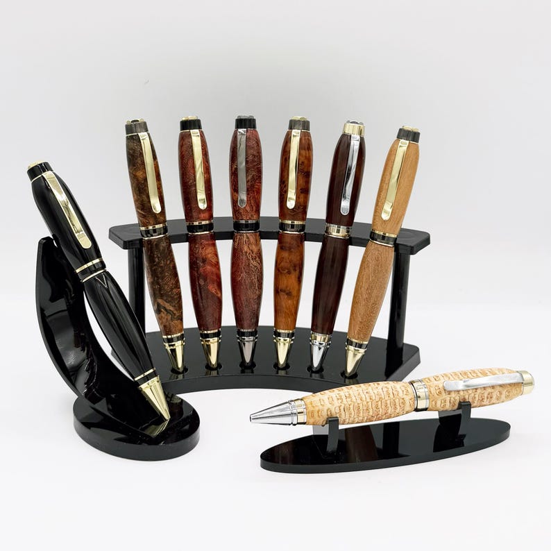 May include: A selection of handcrafted pens with diverse wood grains and finishes, presented on black stands. The pens have gold and silver details, displaying a spectrum of colours from dark brown to light tan. One black pen is also shown.