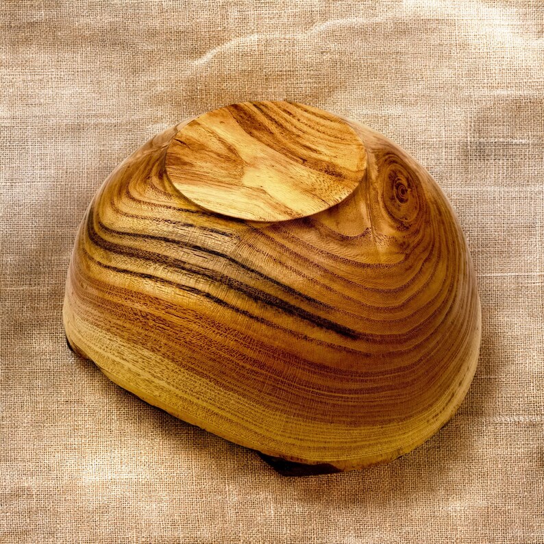 Op de afbeelding: Een houten kom met een natuurlijke afwerking, voorzien van opvallende bruine houtnerven en een ronde voet. De kom staat ondersteboven en toont de gladde, afgeronde buitenkant en de duidelijke patronen van de houtnerf op het oppervlak.
