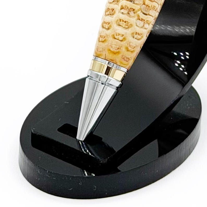May include: A close-up of a pen with a textured, light brown barrel, silver tip, and gold accents. The pen is displayed on a black, oval-shaped stand. The pen's design is elegant and detailed.