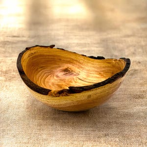 Op de afbeelding: Een houten kom met een natuurlijke levende rand met donkere, ruwe schorsdetails. De binnenkant toont een lichtbruine houtnerf met wervelende patronen en een gladde, gepolijste afwerking, die de organische textuur van het hout benadrukt.