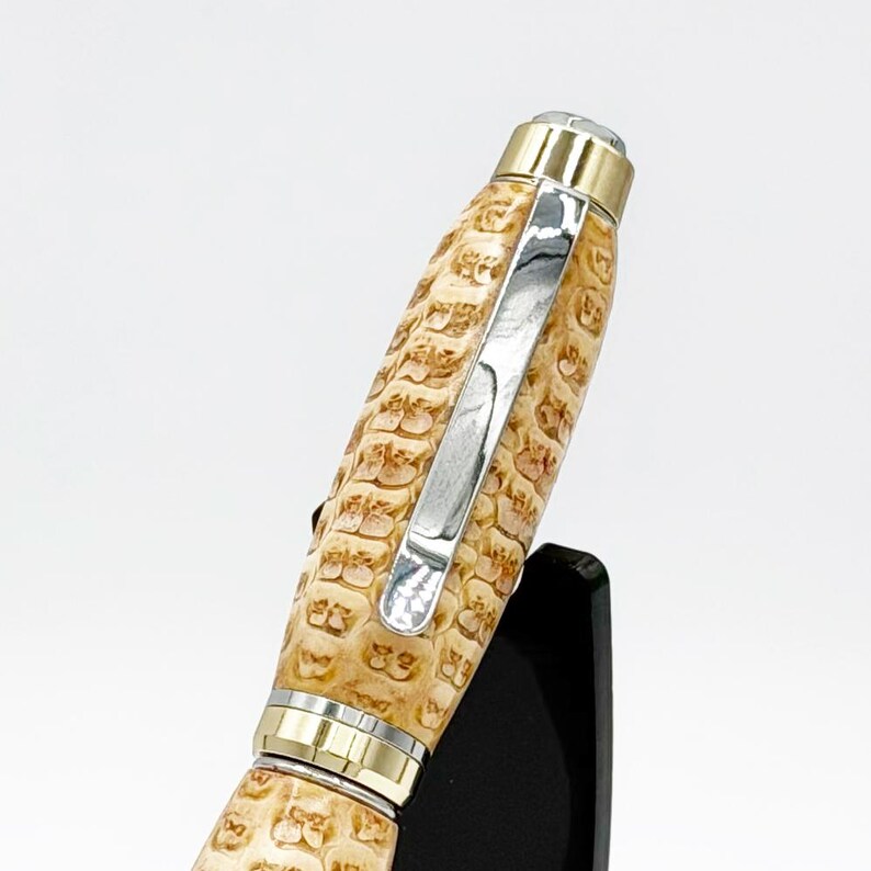 May include: A handcrafted pen with a light brown, textured barrel. The pen features gold and silver-colored metal accents, including a clip and trim. The pen is displayed against a white background, showcasing its intricate design and craftsmanship.