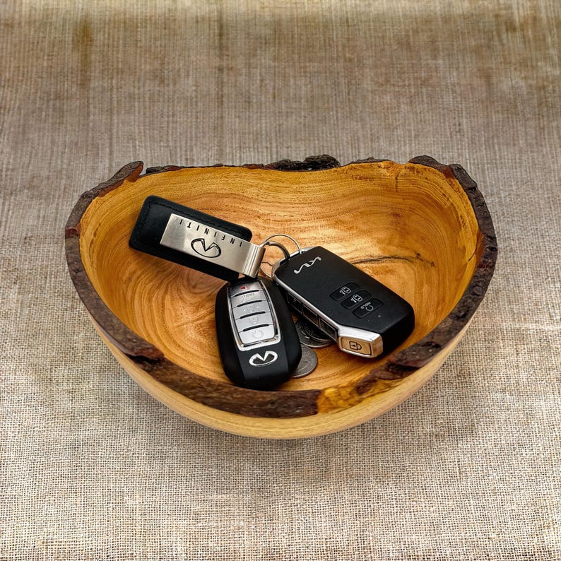 Op de afbeelding: Een houten kom met een natuurlijke rand, een lichtbruine binnenkant en een donkere schorsrand. In de kom liggen twee zwarte autosleutels, &eacute;&eacute;n met een Infiniti-logo en &eacute;&eacute;n met een Kia-logo, samen met een kleine metalen sleutel en zilveren munten.