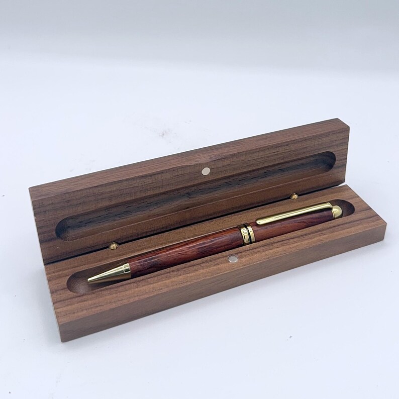 May include: A wooden pen and case. The pen has a dark reddish-brown barrel with gold-coloured accents. The rectangular case has a hinged lid and a magnetic closure. The pen is inside the open case, which is also made of wood.