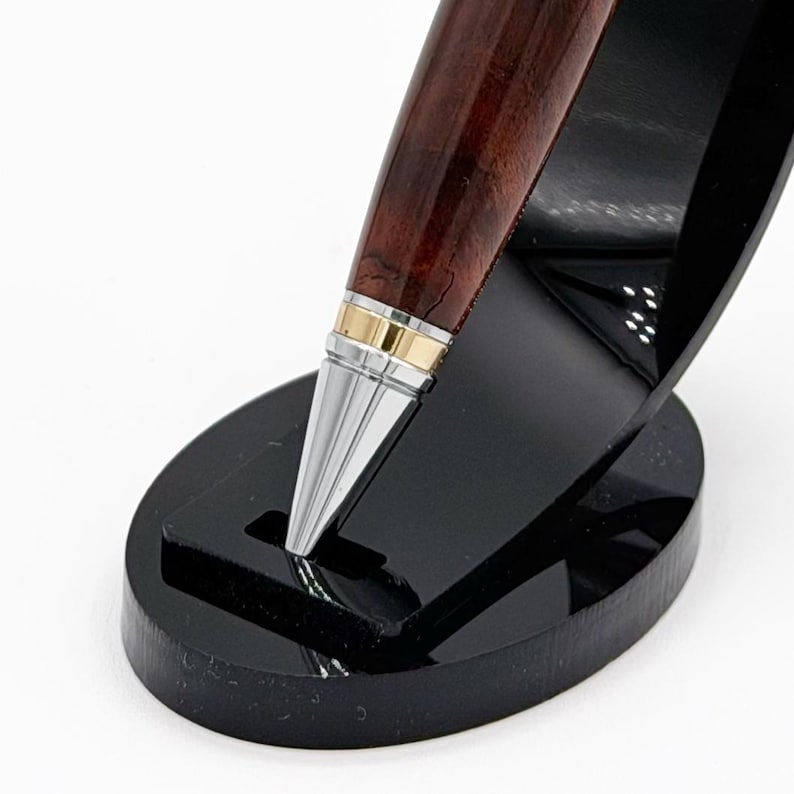 May include: A pen displayed on a black stand. The pen features a dark wood grain barrel with gold and silver detailing. The stand is oval and has a glossy finish.