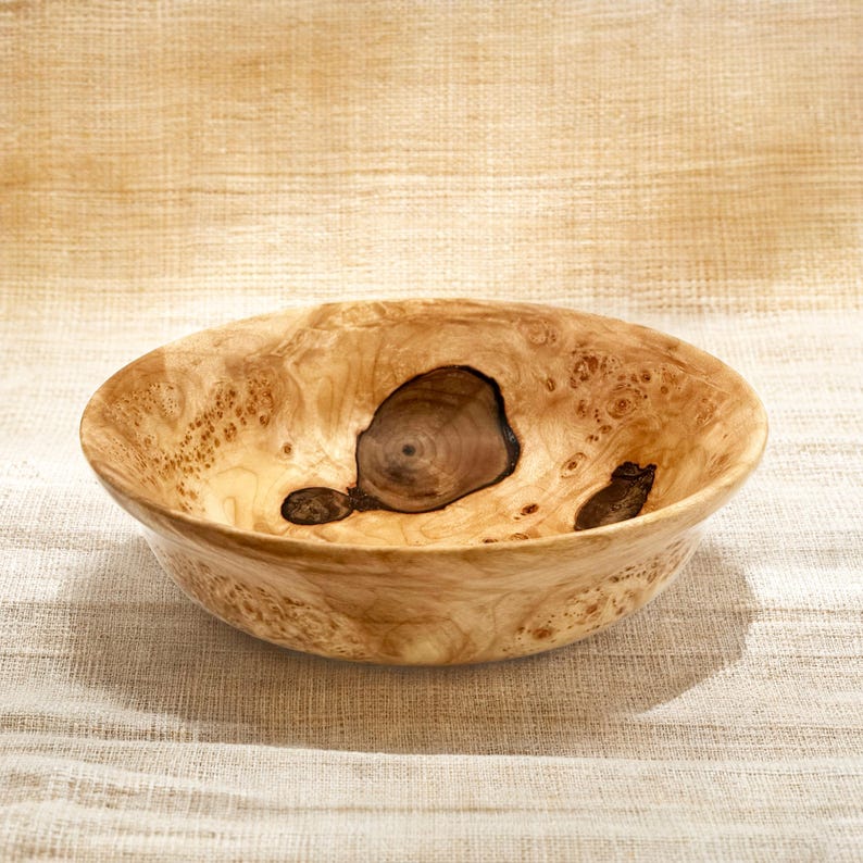 Puede incluir: Un cuenco redondo de madera de ra&iacute;z de color claro, con un acabado liso y pulido. El interior muestra patrones de nudos org&aacute;nicos oscuros y prominentes, junto con texturas de vetas arremolinadas que contrastan con la superficie clara.