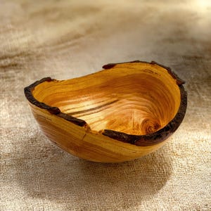 Op de afbeelding: Een handgemaakte houten kom met een natuurlijke levende rand met donkere, getextureerde schorsresten. De binnenkant toont een gepolijste, goudbruine houtnerf met wervelende patronen en een glad, concaaf oppervlak dat de organische vorm benadrukt.