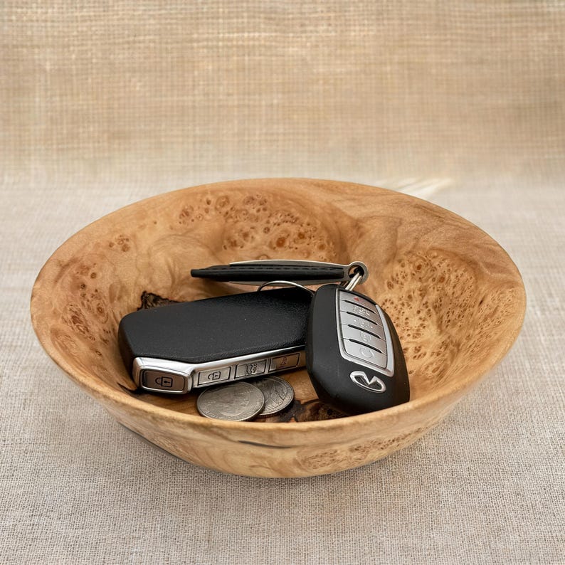 Puede incluir: Un cuenco de madera redondo y poco profundo con un patr&oacute;n de grano de madera de ra&iacute;z natural con texturas arremolinadas en marr&oacute;n claro y oscuro. Dentro del cuenco hay dos llaves de coche negras con botones plateados y dos monedas plateadas.