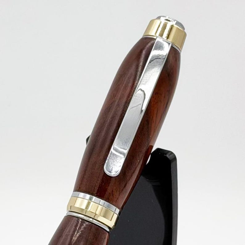 May include: A wooden pen with a silver clip and gold detailing. The pen has a dark brown, polished finish and a rounded shape. The pen is displayed on a black stand against a white background.