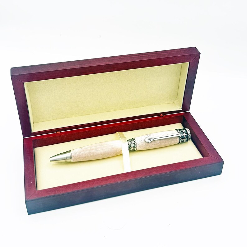 May include: A wooden pen with a light-colored barrel and silver accents rests inside a burgundy wooden presentation box with a cream-colored interior. The pen has a silver tip and a decorative silver band near the top.