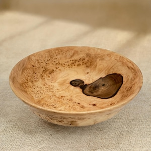Puede incluir: Un cuenco de madera redondo con una veta natural de color tostado claro y una prominente inclusi&oacute;n de nudo irregular de color marr&oacute;n oscuro en el centro. La superficie muestra un patr&oacute;n moteado y texturizado en el interior.