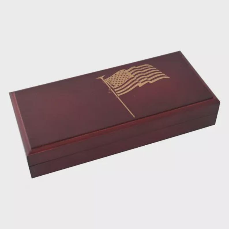May include: A rectangular, burgundy-colored wooden box with a hinged lid. The lid features a gold-colored engraving of the American flag. The box has a polished finish and appears to be designed for storage or display.