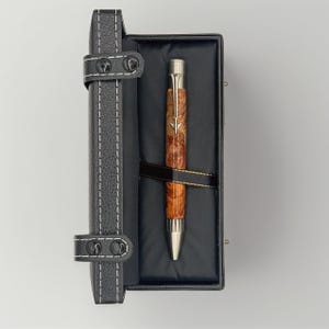 May include: A wooden pen with a silver tip and accents rests in a black leather case. The pen features a brown, burl wood barrel and a silver arrow design. The case is black with white stitching and a leather strap closure.