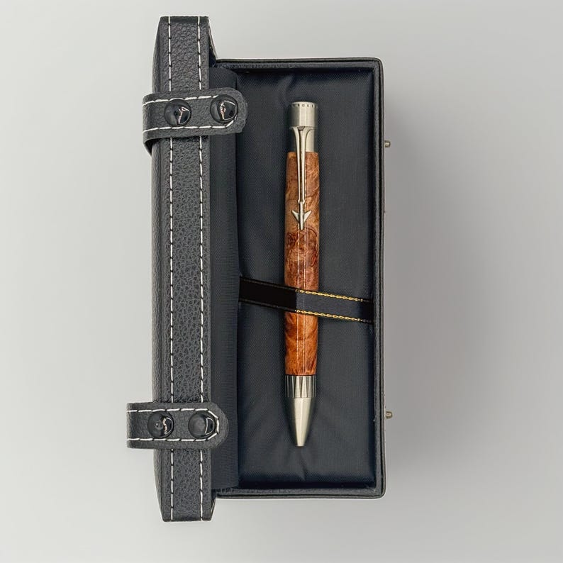 May include: A wooden pen with a silver tip and accents rests in a black leather case. The pen features a brown, burl wood barrel and a silver arrow design. The case is black with white stitching and a leather strap closure.