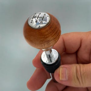 May include: A wooden bottle stopper with a chrome top, featuring a gear shift pattern. The wooden sphere is brown, and the chrome top displays the numbers 1, 2, 3, 4, 5, and R. The stopper is black.