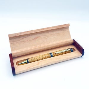 May include: A wooden pen with a gold-colored tip and accents rests inside a light-colored wooden box with a dark red border. The pen has a patterned design and a gold clip.