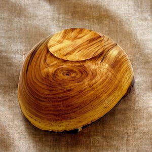 Op de afbeelding: Een ronde houten kom met een opvallend natuurlijk nerfpatroon in wervelende licht- en donkerbruine tinten. De kom heeft een glad, gepolijst oppervlak en een duidelijke ronde voet, die de organische textuur en de ringen van het hout laat zien.