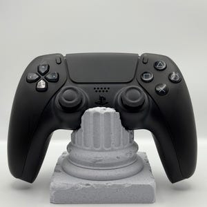 May include: A black video game controller rests on a gray, textured pedestal. The controller has various buttons and joysticks. The text "Free & Fast Shipping!" is at the top of the image.