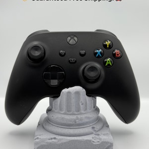 May include: A black Xbox controller rests on a gray, textured, column-shaped stand. The controller has colored buttons and a directional pad. The text "Guaranteed Free Shipping!" is at the top.